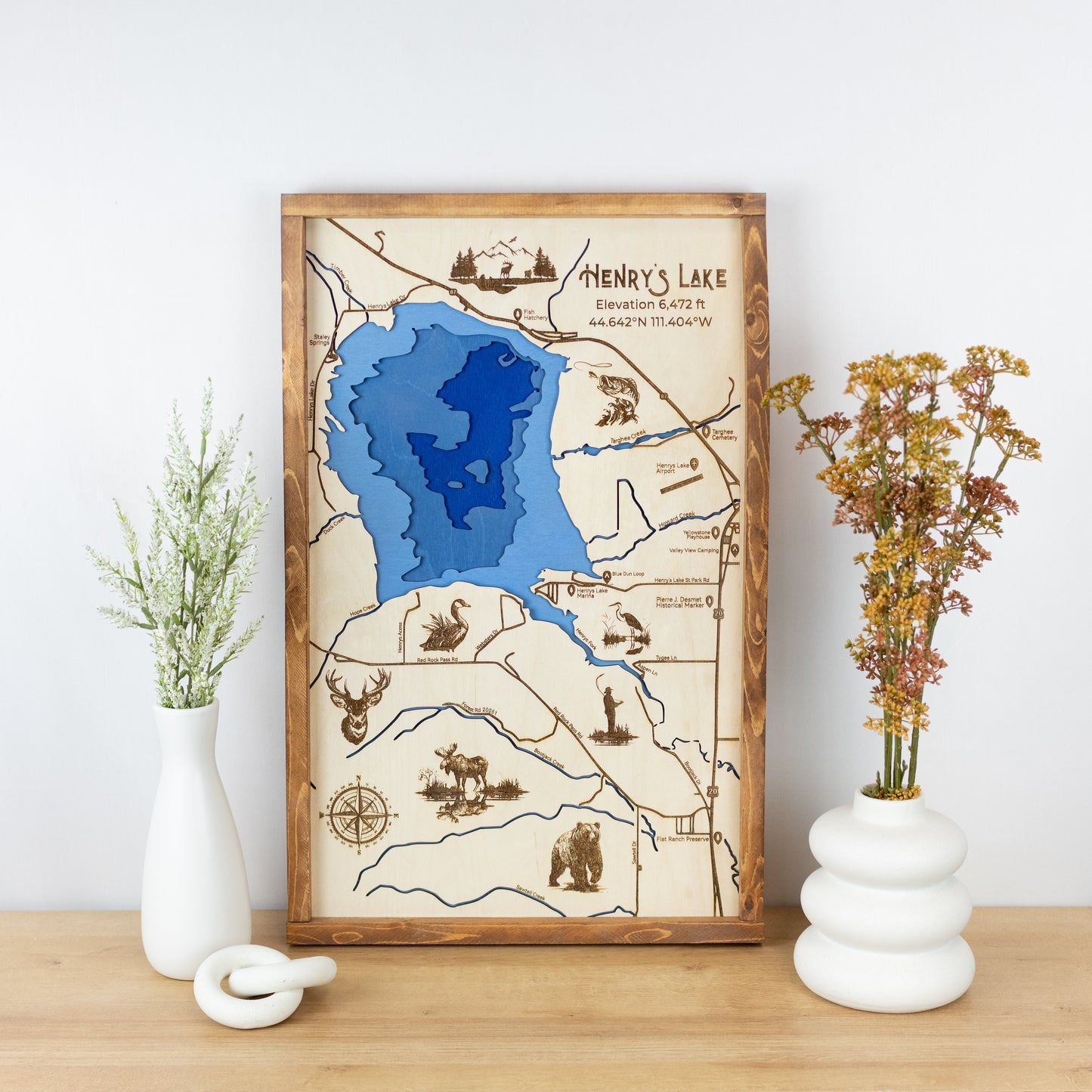 Laser-engraved wooden map of Henry's Lake with layered blue inlay, framed and styled on a tabletop with floral vases.