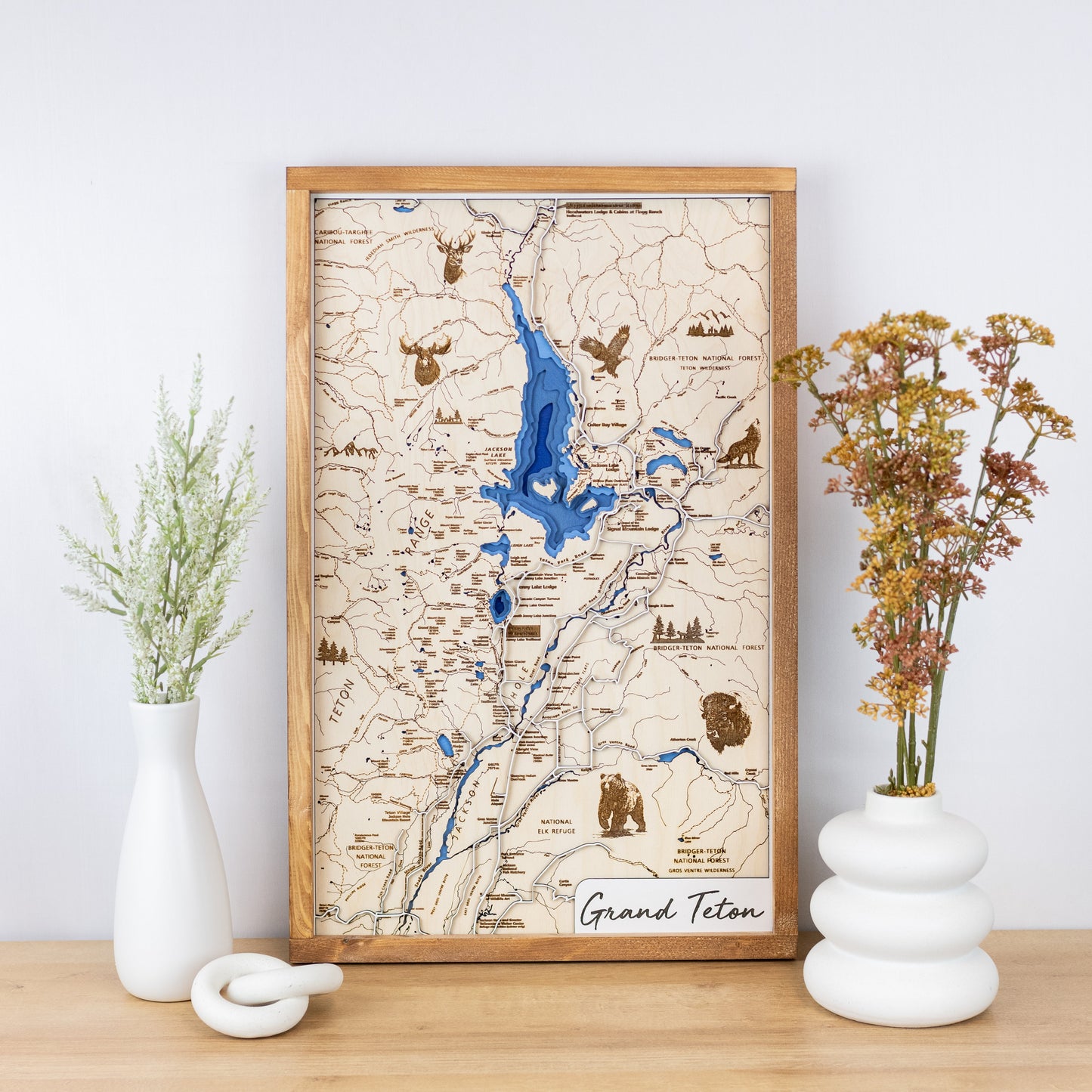 Laser-engraved wooden map of Grand Teton National Park with layered blue inlay and trail details framed and styled on a tabletop with floral vases.