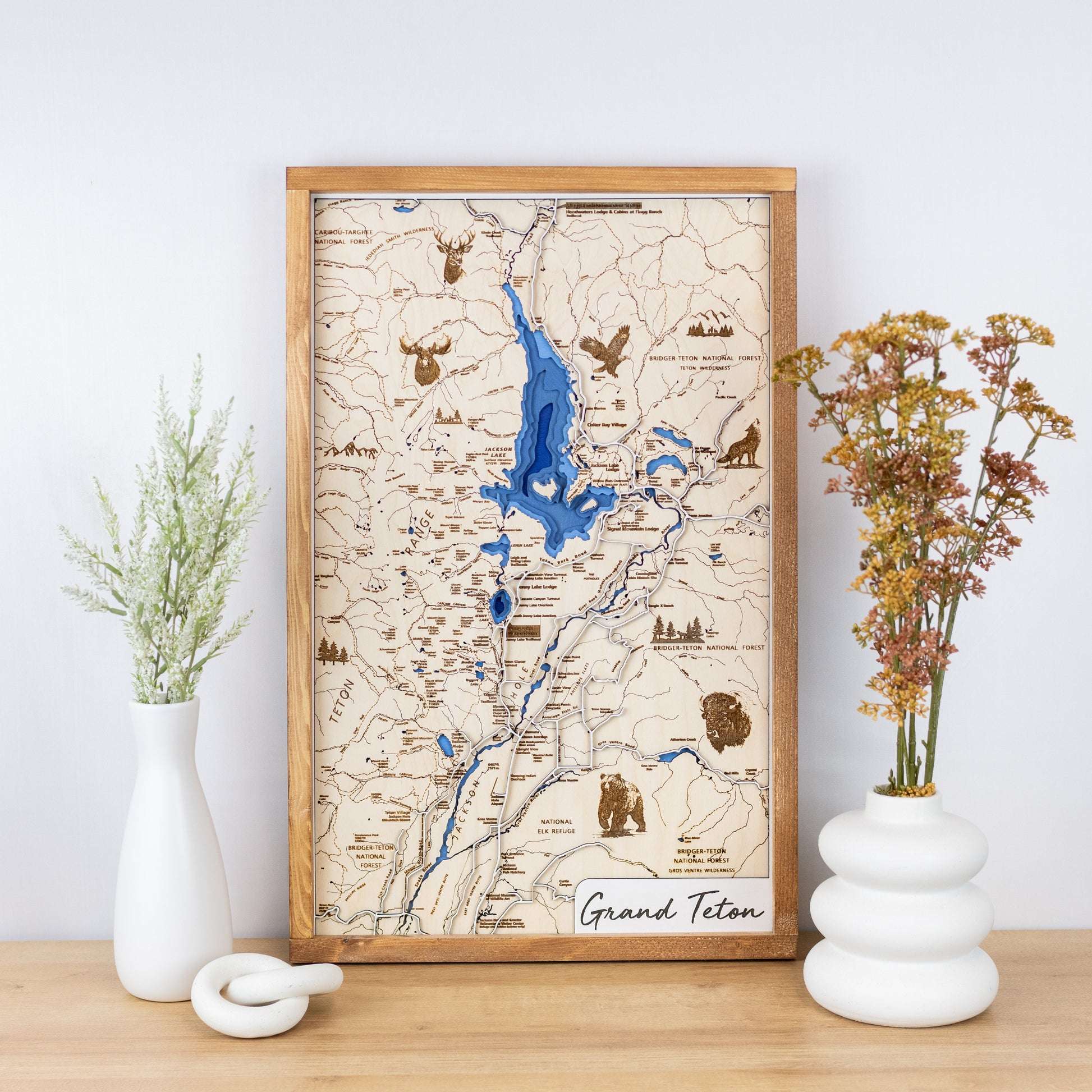 Laser-engraved wooden map of Grand Teton National Park with layered blue inlay and trail details framed and styled on a tabletop with floral vases.