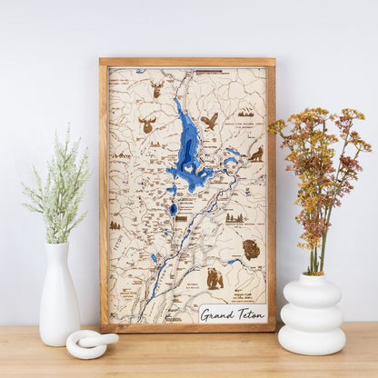 Laser-engraved wooden map of Grand Teton National Park with layered blue inlay and trail details framed and styled on a tabletop with floral vases.